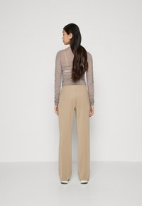 Top cu mânecă lungă, transparent, în nuanță de taupe, cu textură pliată și pantaloni largi bej. Modelul este cu spatele, evidențiind designul și croiul din spate.