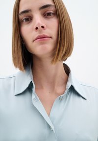 Cămașă albastră deschis, cu guler, având o textură netedă. Prezintă un sistem de închidere cu nasturi în față și detalii de cusut subtile pe linia gulerului.
