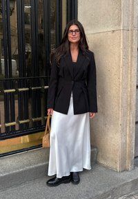 Blazer noir ajusté sur un haut noir près du corps et une jupe fluide en satin blanc, associé à des bottes noires et un sac à main marron.