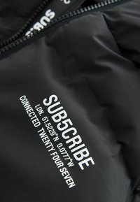Tissu noir avec du texte imprimé en blanc disant "SUB5CRIBE LDN. 51.5229°N 0.0777°W CONNECTED TWENTY FOUR SEVEN" près d'une fermeture éclair.
