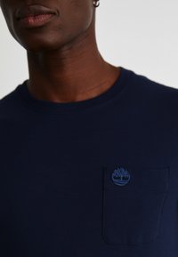 Tricou din bumbac de culoare bleu marin, cu guler rotund, dotat cu un mic logo brodat cu un copac deasupra unui buzunar aplicat. Textură netedă, design minimalist.