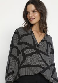 Bluza u crno-bijelom uzorku s V-izrezom, koja se ističe hrabrim geometrijskim linijama i opuštenim krojem. Izrađena od lagane tkanine s dugim rukavima.