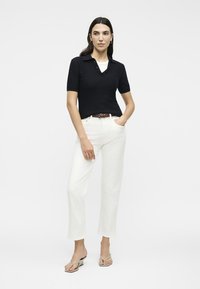 Γυναίκα στέκεται φορόντας μαύρο κοντομάνικο πουκάμισο με γιακά, λευκό παντελόνι με ψηλή μέση, καφέ ζώνη και μπεζ σανδάλια με τακούνι σε λευκό φόντο.