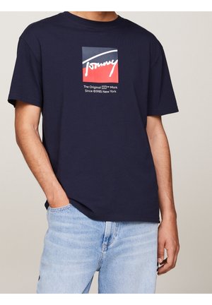 Man wearing navy blue Tommy Hilfiger t-shirt with logo and text, paired with light blue jeans, hand in pocket.