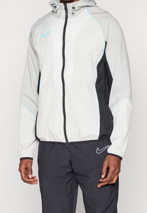 Man draagt een witte en zwarte Nike capuchonjas met een rits aan de voorkant en een zwarte broek met een wit Nike-logo op de linker dij.