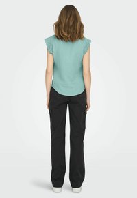 Blusa verde menta con orli in pizzo a forma di scallop, abbinata a pantaloni cargo neri. La blusa presenta un orlo arrotondato e un tessuto testurizzato.