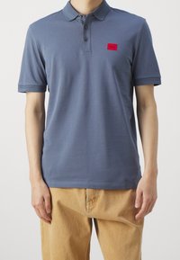 Man med blå kortärmad pikétröja med röd Hugo-logotyp på bröstet och beige byxor, stående med armarna avslappnat vid sidorna.