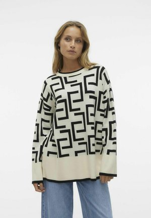 Übergroßer cremefarbener Pullover mit schwarzen geometrischen Mustern, geripptem Halsausschnitt und Bündchen sowie einer weichen Textur. Kombiniert mit hellblauen Jeans.
