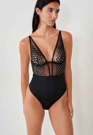 Maillot de bain noir avec des panneaux en maille transparente présentant des motifs circulaires texturés, un décolleté en V profond et des jambes hautes. Bretelles ajustables.
