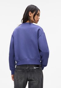 Purple cotton sweatshirt with a crew neck and ribbed hem, paired with dark jeans featuring a logo patch on the back pocket.