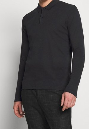Polo noir à manches longues avec un col et une patte de boutonnage à deux boutons. Tissu doux avec une texture lisse. Porté avec un pantalon à carreaux.