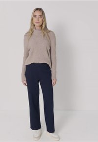 Vrouw staat in een beige coltrui, donkerblauwe wijde broek en witte sneakers tegen een effen lichte achtergrond.