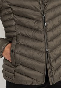 Fodrad jacka i mörk olivgrön, med dragkedjestängning, horisontellt quiltad design och sidofickor med knappdetaljer.