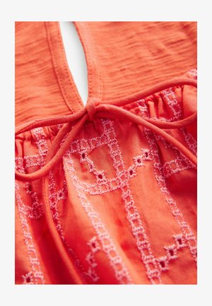 Top en tissu corail avec un décolleté en forme de trou de serrure et un détail de lien froncé. Broderies florales en rose clair ajoutent texture et intérêt visuel.