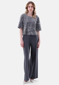 Silberner, mit Pailletten besetzter Cropped-Top mit kurzen Ärmeln, kombiniert mit dunkelgrauen, weit geschnittenen Hosen und weißen High Heels. Schlichter Ausschnitt und Passform.