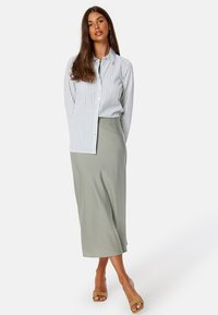 Camicia bianca con bottoni, a righe verticali grigie, abbinata a una gonna verde chiaro di lunghezza media. Modella che indossa sandali beige. Texture morbide, vestibilità rilassata.