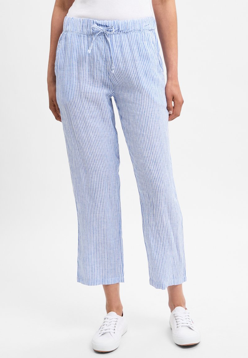 Femme portant un pantalon à cordon bleu clair et blanc à rayures verticales et des baskets blanches, debout devant un fond uni.