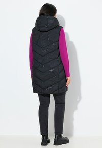 Žena v černé vzorované polstrované vestě s kapucí, magentovém svetru s dlouhým rukávem, černých džínách a černých kotních botách, stojící u bílé zdi.