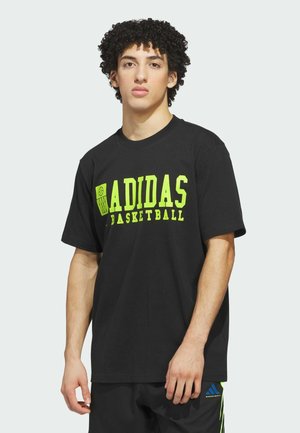 Joven con cabello rizado que lleva una camiseta negra de baloncesto Adidas con texto en neón amarillo y pantalones cortos negros a juego con detalles en azul y verde.