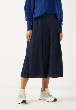 Personne portant une jupe midi en jean foncé, un pull bleu et des baskets blanches, debout sur un fond clair uni.