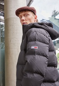Crna puffer jakna s patentnim zatvaračem sprijeda i kapuljačom, koja sadrži kvadratne šavove i logotip na rukavu. Teksturovana vunena kapa.