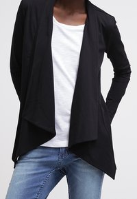 Svart långärmad cardigan med öppen framsida, tillverkad av mjukt tyg. Kombineras med en vit T-shirt och blå jeans. Slät struktur.