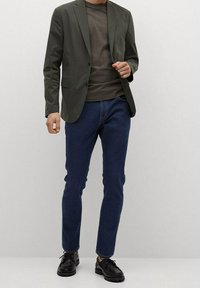 Blazer verde oscuro sobre una camisa gris, combinado con jeans oscuros ajustados y zapatos negros con cordones. Tela suave con un diseño estructurado.