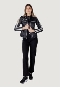 Giacca leggera nera con strisce bianche e toppe ricamate; presenta una chiusura a zip e un design aderente, abbinata a pantaloni neri e sneaker.