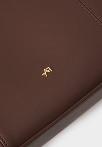Brun læderportefølje med glat tekstur og synlige syninger. Guld emblem vist i det nederste hjørne. Enkel, rektangulær form.