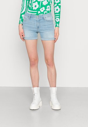 Calções de ganga azul claro com bainha dobrada, combinados com botas de combate brancas com atacadores. A parte superior apresenta uma blusa de manga longa com padrão floral verde.