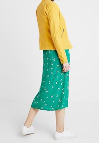 Giacca in pelle gialla con dettagli trapuntati, abbinata a una gonna midi floreale verde. La modella indossa sneaker bianche, mostrando un look casual.