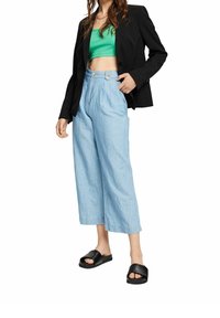 Femme portant un crop top vert, un blazer noir, un jean bleu clair taille haute à jambes larges, et des sandales noires, debout avec une main dans la poche.