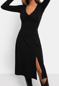 Femme portant une robe noire à manches longues avec un col en V et une fente sur le côté, assortie à des bottes noires montantes, posant avec les bras levés.