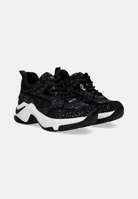 Zapatillas negras con un upper texturizado que presenta piedras brillantes, cordones negros y una entresuela blanca. Diseño de suela curva con una banda de rodadura de goma negra.
