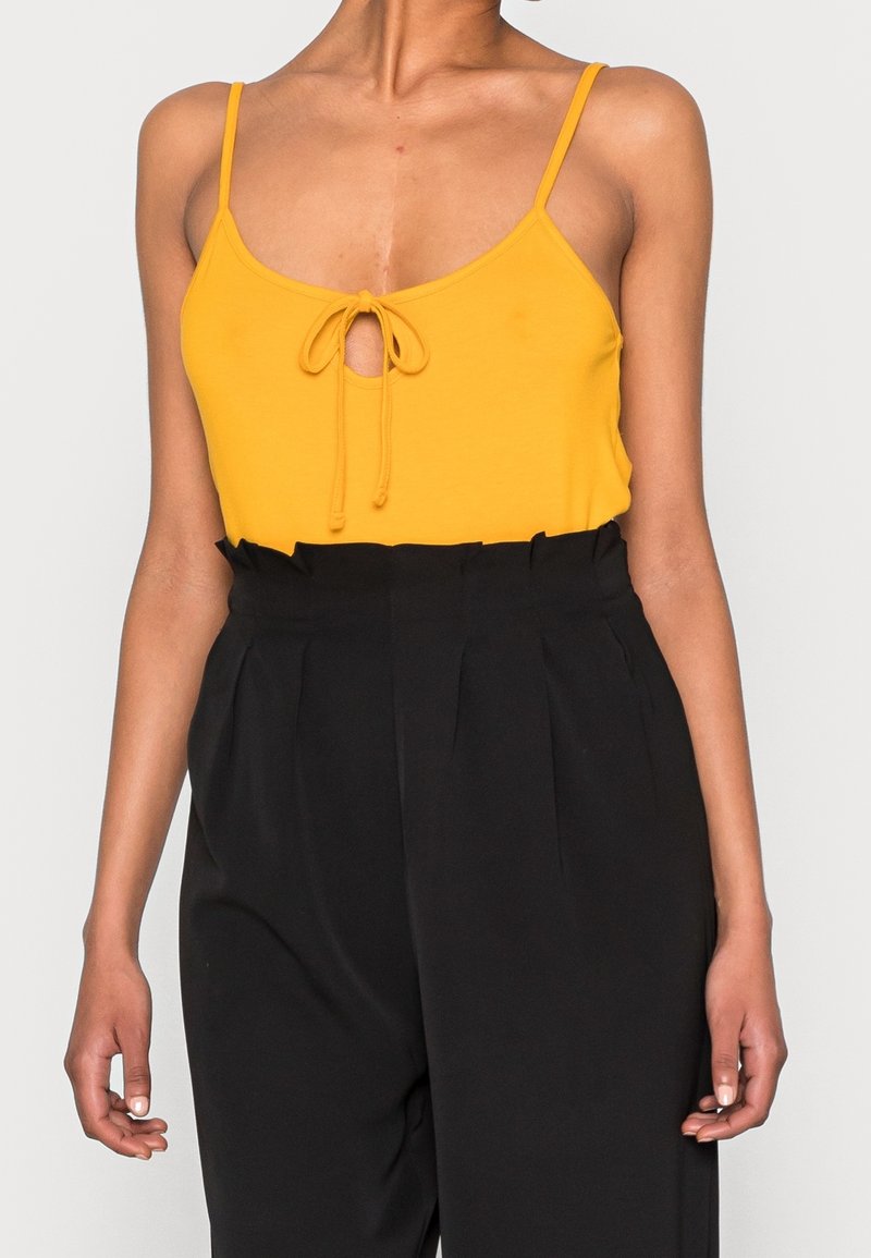 Gul ärmlös topp med en nyckelhål och knytdetalj framtill, i kombination med högmidjade svarta byxor. Slät textur, avslappnad design.