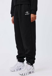 Zwarte trainingsbroek met een elastische tailleband en manchetten, met een wit "PXP" logo op de linker bovenbeen. Gepaard met witte sportschoenen.