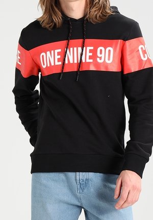 Mężczyzna w czarnej bluzie z kapturem z czerwonym paskiem na klatce piersiowej oraz białym napisem "ONE NINE 90", ubrany w jasnoniebieskie dżinsy.