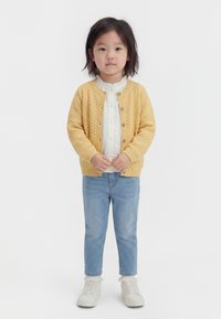 Noor laps keskmise pikkusega tumedate juustega, kes kannab kollast kootud kardigani, valget pluusi, helerohelisi teksaseid, valgeid sokke ja valgeid jalanõusid.