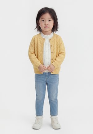 Noor laps keskmise pikkusega tumedate juustega, kes kannab kollast kootud kardigani, valget pluusi, helerohelisi teksaseid, valgeid sokke ja valgeid jalanõusid.