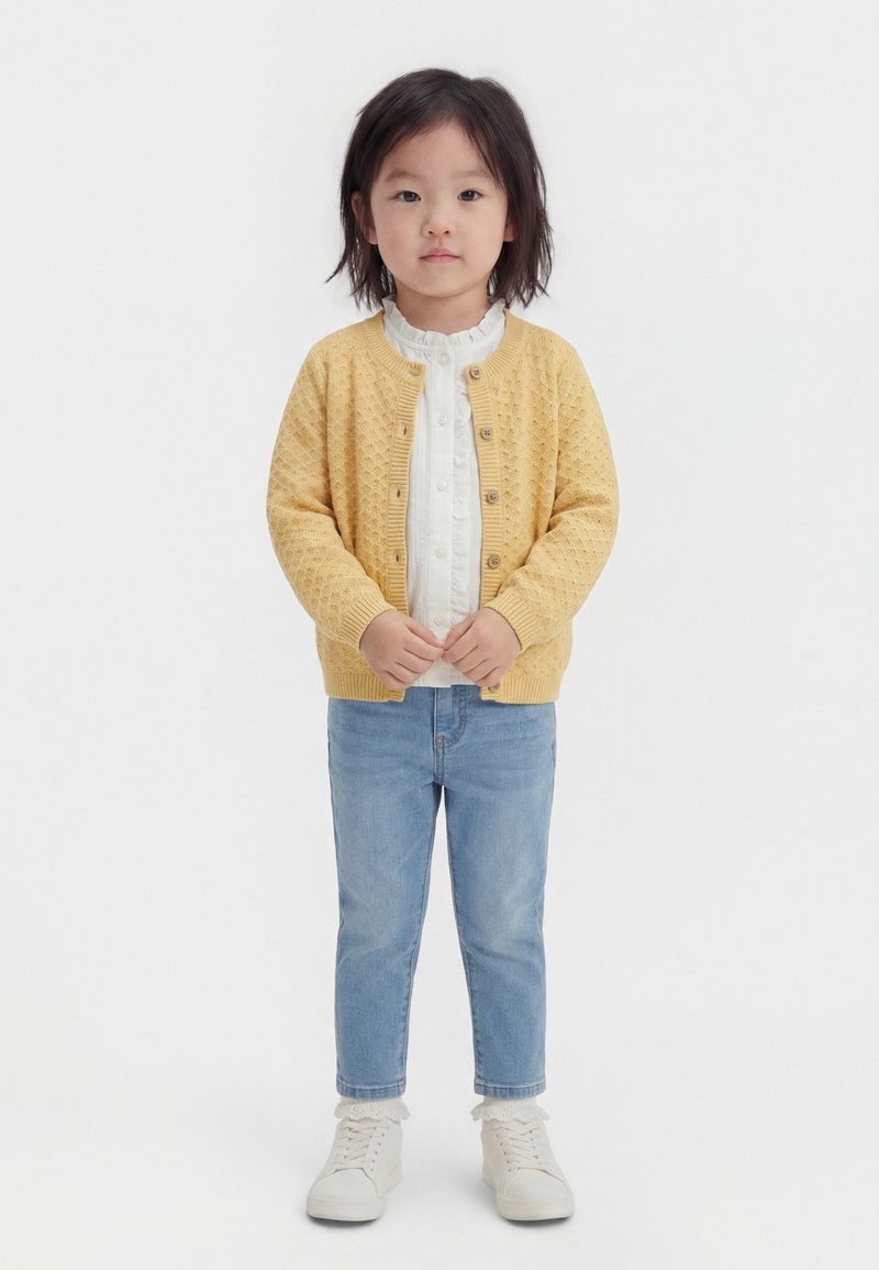 Noor laps keskmise pikkusega tumedate juustega, kes kannab kollast kootud kardigani, valget pluusi, helerohelisi teksaseid, valgeid sokke ja valgeid jalanõusid.