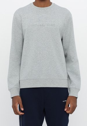 Person trägt ein hellgraues Sweatshirt mit langen Ärmeln und "MICHAEL KORS" geprägt auf der Brust sowie dunkelblaue Hose mit kleinem Logo.