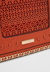 Czerwona perforowana skórzana torebka z akcentem w postaci złotego łańcuszka i złotą plakietką z logo. Gładka faktura z geometrycznymi wycięciami.