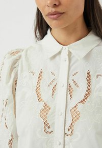 Hvit bluse med blomsterbroderi, puffete blondermerker, klassisk krage og knappelukking, med utskjæringer gjennom hele stoffet.