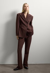 Costume marron sur mesure comprenant un blazer structuré à deux boutons et un pantalon à jambe droite. Le tissu semble lisse avec une texture minimale.