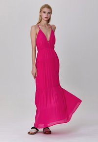 Robe maxi rose avec des bretelles spaghetti, décolleté en V profond, taille froncée et jupe à volants. Le mannequin porte des sandales plates. Tissu léger avec un léger éclat.