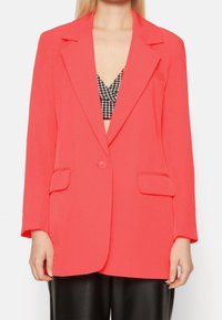 Blazer rosa corallo realizzato in tessuto morbido, con colletto a pistagna, chiusura con un bottone e due tasche frontali.
