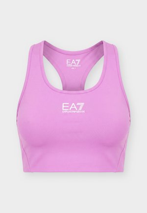 Jeune femme debout portant un soutien-gorge de sport rose, une jupe athlétique noire, des chaussettes blanches et des baskets blanches sur un fond uni.