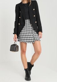 Vrouw in een zwarte blazer met gouden knopen, zwart top, zwart-wit geruit rokje, zwarte veterschoenen, en een gestikte zwarte handtas.