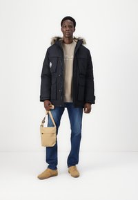 Giacca nera imbottita con cappuccio rifinito in pelliccia, felpa beige, jeans blu e sneakers color beige. Porta una borsa a tracolla beige.