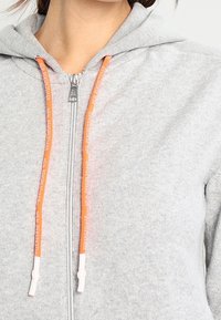 Szara bluza z kapturem zapinana na zamek, o miękkiej fakturze, z pomarańczowym sznurkiem zakończonym białymi końcówkami oraz zamkiem przy szyi.